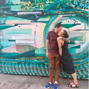 A man and a woman hugging standing in front of a very colorful wall 