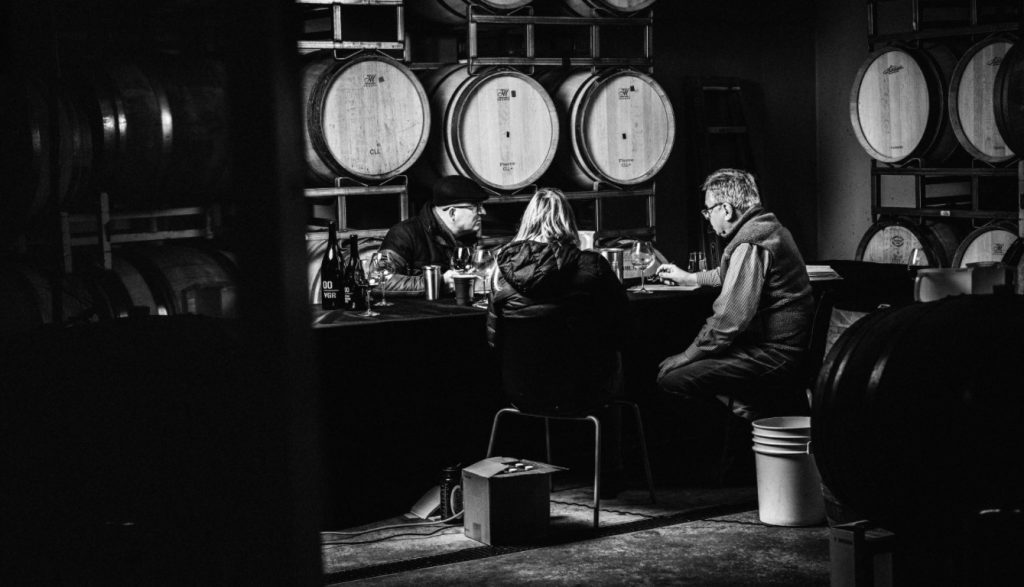 Black and white photo with three people sitting around a table in a barrel room tasting wine