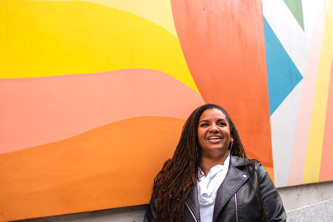 A smiling woman leans against a brightly painted wall
