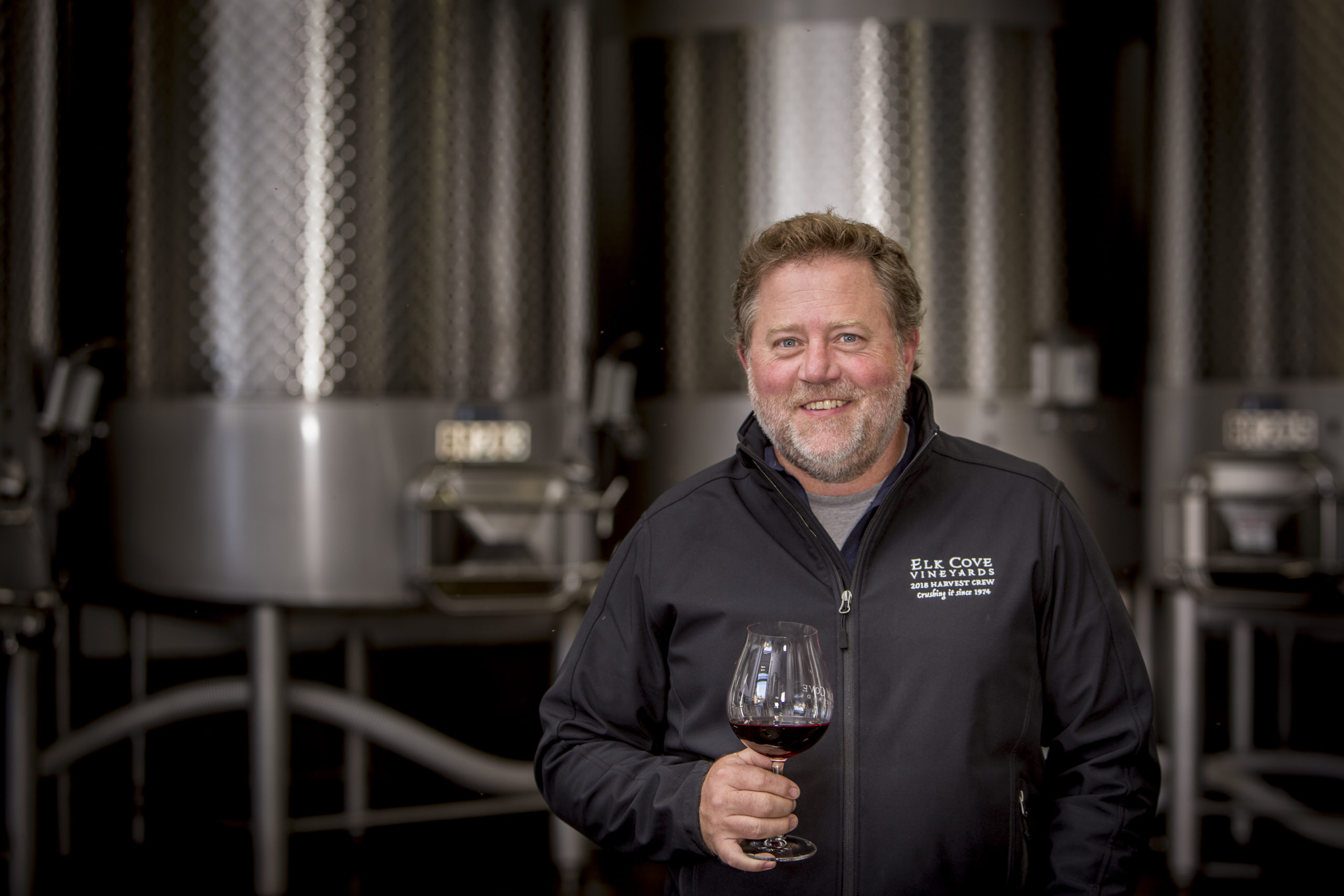 winemaker Adam Campbell smiling with glass of red wine in front of fermentation tanks