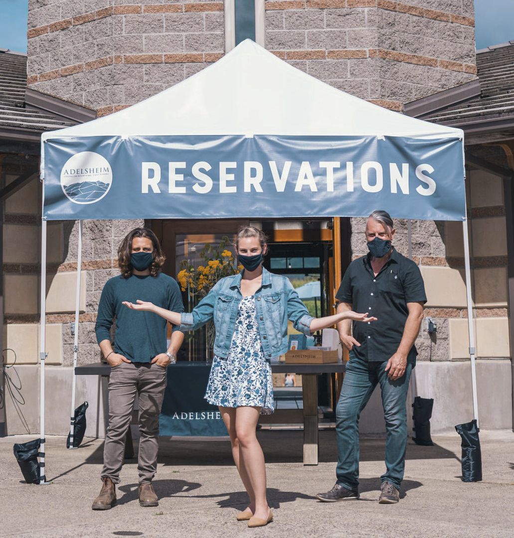 One woman and two men standing in front of a small tent outdoors that reads "reservations" while wearing COVID face coverings