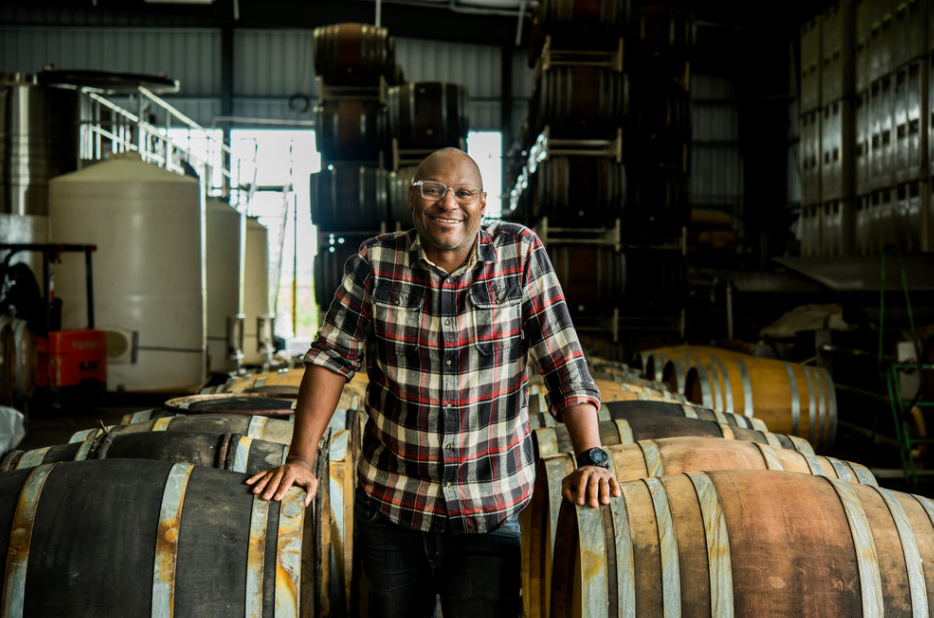 Winemaker Andre Mack stands smiling inside a wine cellar, between rows of wine barrels