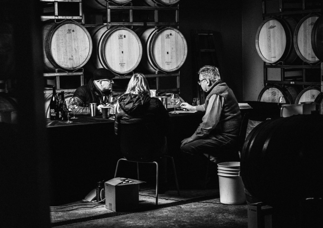 Black and white photo of three people sitting in a barrel room at a table tasting wine