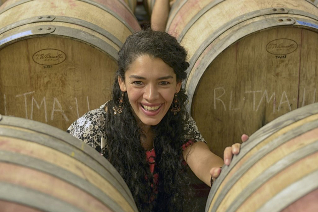 A woman kneels between wooden wine barrels while smiling at the camera