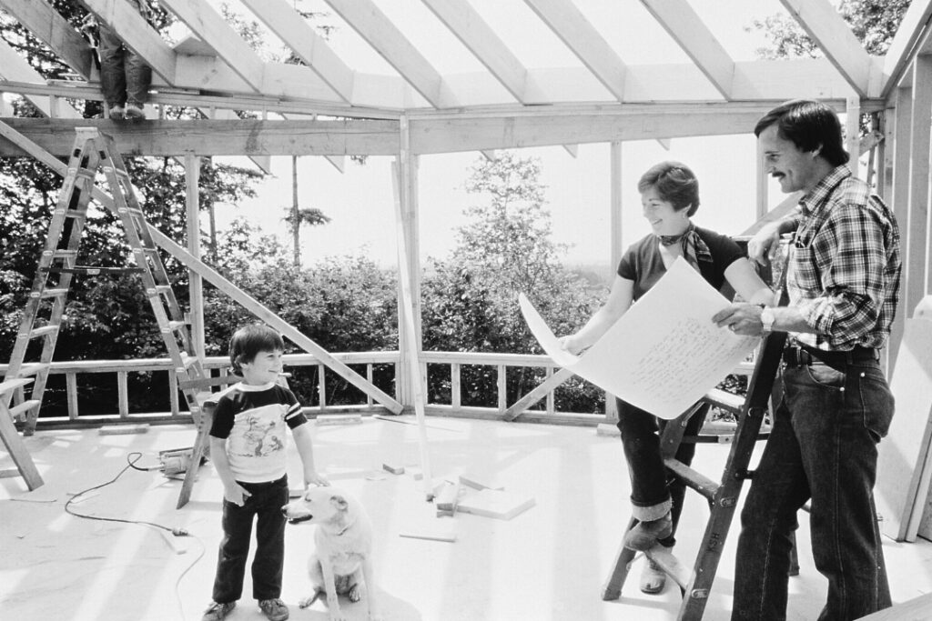 Two adults stand holding a large paper between them while smiling at a child. All three stand inside a building being built with only timber framing completed.