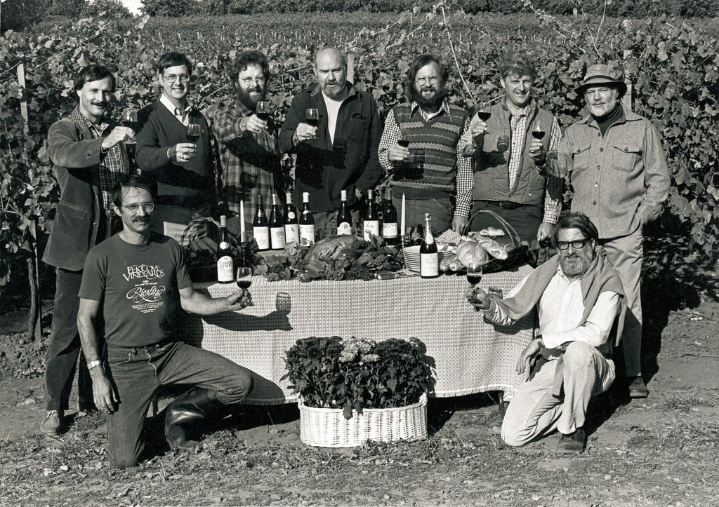A large group of people stand around a table in a vineyard decorated with Thanksgiving foods and many wine bottles.