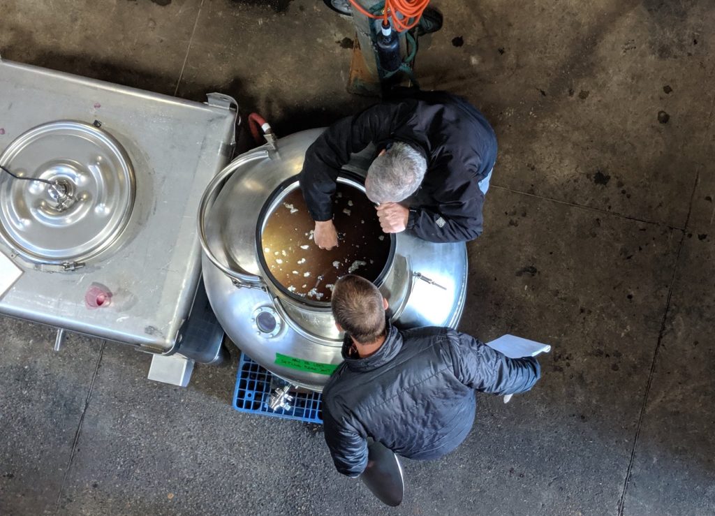 A photograph overhead of two people looking into a large vessel filled with wine.