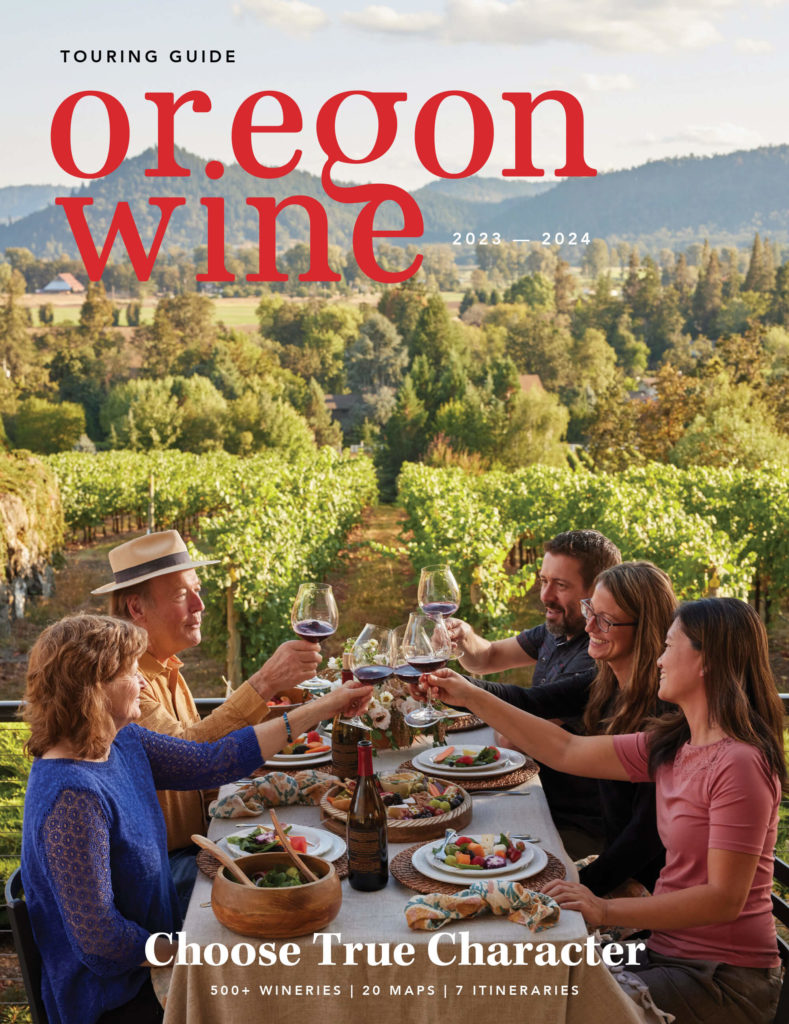 An image of the cover of the 2023-24 Oregon Wine Touring Guide, featuring 5 adults sitting at an outdoor table, eating a meal, toasting cheers with glasses of red wine, with vineyards in the background.